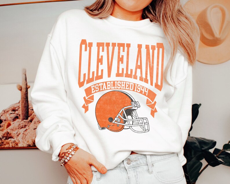 以下が含まれることがあります： オレンジ色のブロック文字で「CLEVELAND」、その下に「ESTABLISHED 1944」とフットボールのヘルメットのグラフィックがプリントされた白いスウェットシャツ。クルーネックのスタイルです。