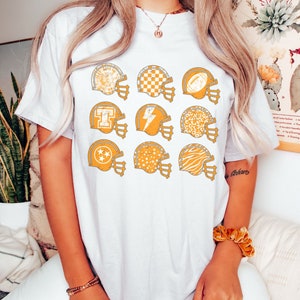 May include: White t-shirt with a distressed look featuring nine yellow football helmets with different patterns, including stars, checkers, and animal prints.