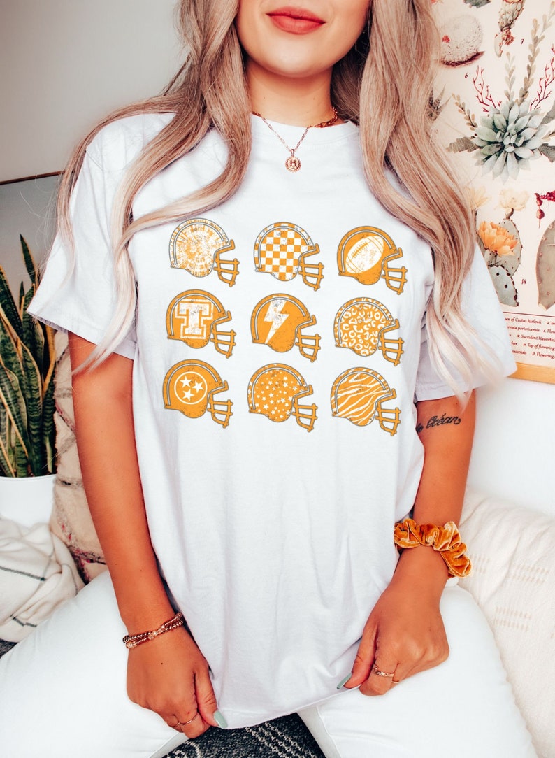 May include: White t-shirt with a distressed look featuring nine yellow football helmets with different patterns, including stars, checkers, and animal prints.