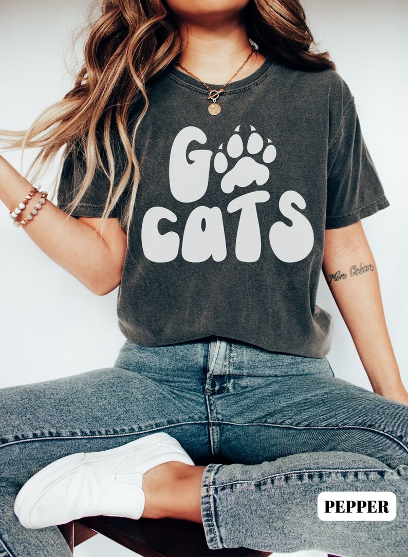 May include: A dark gray t-shirt with a white graphic that says "Go Cats" with a paw print in the "O". The model is wearing blue jeans and white sneakers.