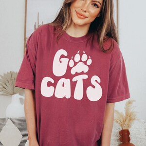 May include: A maroon t-shirt with a white graphic that says "Go Cats" with a paw print in the middle of the "O".