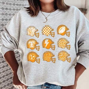 May include: A grey sweatshirt featuring a graphic of nine orange football helmets with various patterns. The patterns include stars, chequered designs, lightning bolts, and animal prints.
