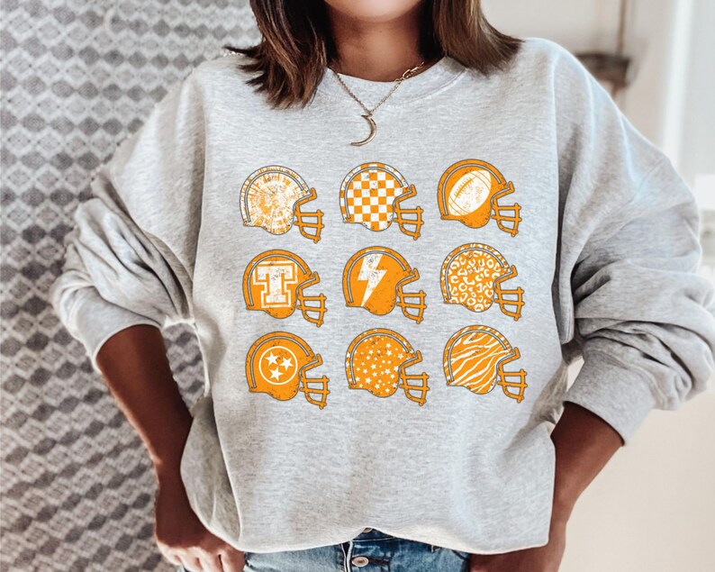 May include: A grey sweatshirt featuring a graphic of nine orange football helmets with various patterns. The patterns include stars, chequered designs, lightning bolts, and animal prints.