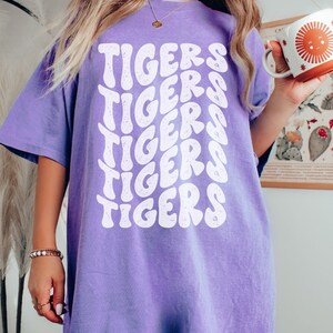 Peut inclure: Un t-shirt violet avec le mot "TIGERS" r&eacute;p&eacute;t&eacute; dans une police r&eacute;tro blanche.