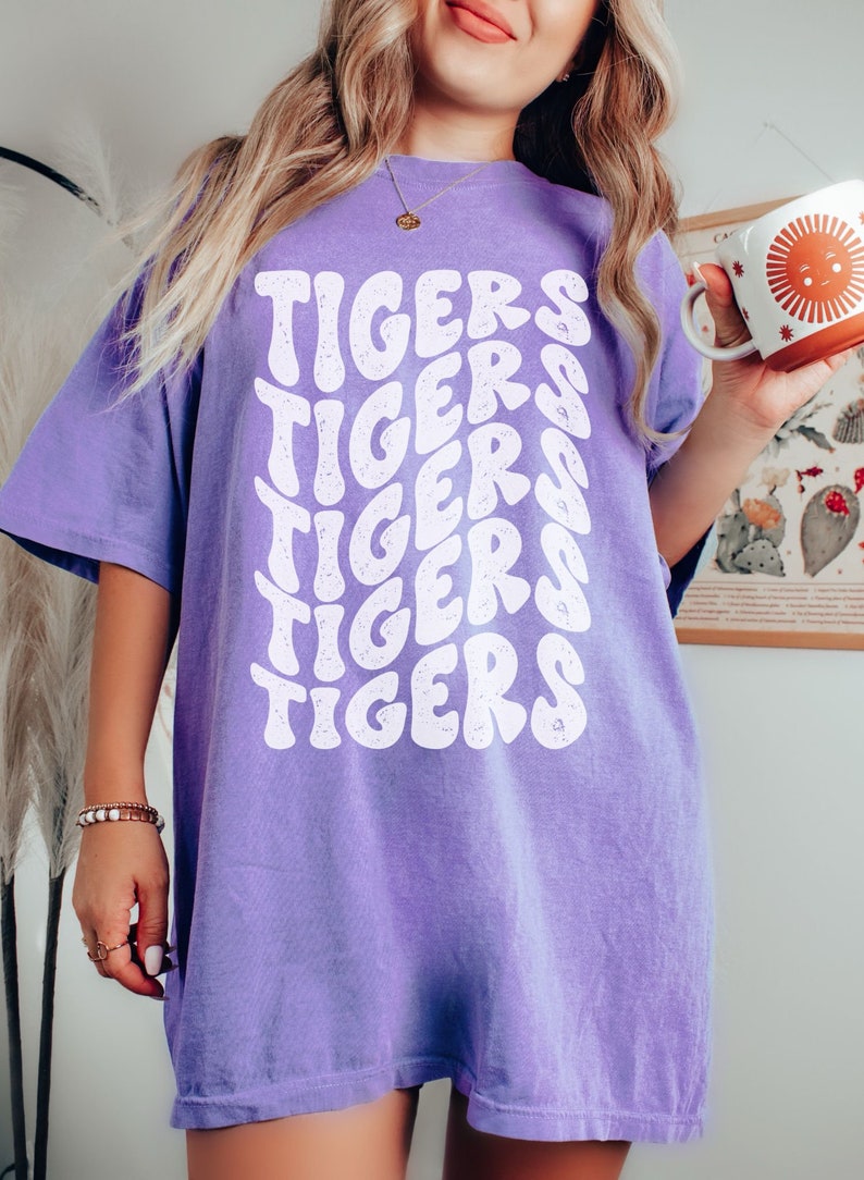 Peut inclure: Un t-shirt violet avec le mot "TIGERS" r&eacute;p&eacute;t&eacute; dans une police r&eacute;tro blanche.