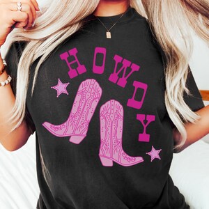 Pode incluir: Camiseta preta com um gr&aacute;fico rosa de botas de cowboy e a palavra "HOWDY" em letras rosas.