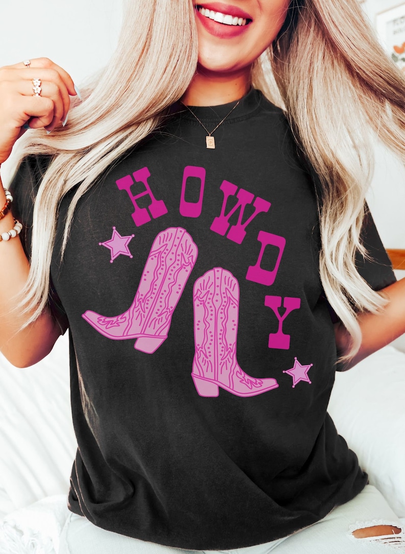 Pode incluir: Camiseta preta com um gr&aacute;fico rosa de botas de cowboy e a palavra "HOWDY" em letras rosas.