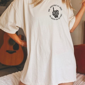 Op de afbeelding: Een wit oversized T-shirt met een zwarte afbeelding van een skelet hand die de middelvinger opsteekt. De afbeelding is omgeven door een cirkel met de tekst "Mrs. Martinez est. 2023 Til Death Do Us Part."