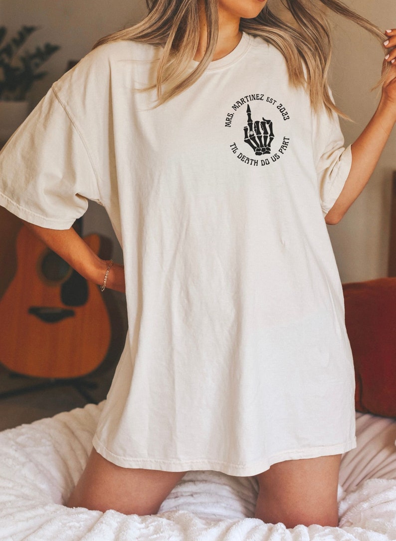 Op de afbeelding: Een wit oversized T-shirt met een zwarte afbeelding van een skelet hand die de middelvinger opsteekt. De afbeelding is omgeven door een cirkel met de tekst "Mrs. Martinez est. 2023 Til Death Do Us Part."