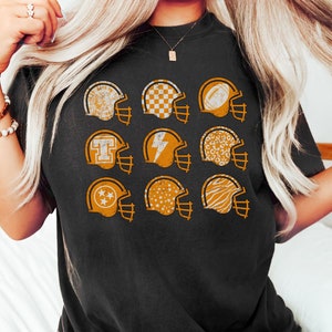 May include: A black t-shirt with a graphic of nine orange football helmets. Each helmet has a different pattern inside, including stars, checkered, and animal print.