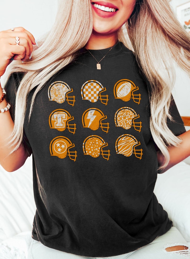 May include: A black t-shirt with a graphic of nine orange football helmets. Each helmet has a different pattern inside, including stars, checkered, and animal print.