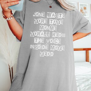 May include: Gray oversized t-shirt with white text that reads "Jone Waste Yore Toye Monme Yore All Red The Voice Inside Moye Yedd"