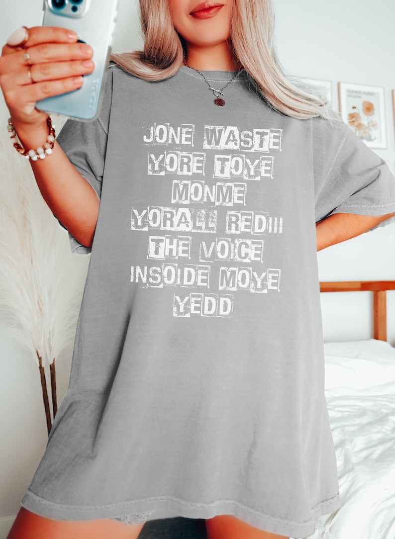 May include: Gray oversized t-shirt with white text that reads "Jone Waste Yore Toye Monme Yore All Red The Voice Inside Moye Yedd"