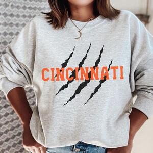 May include: A grey sweatshirt with a black claw mark graphic and the word "CINCINNATI" in orange lettering.