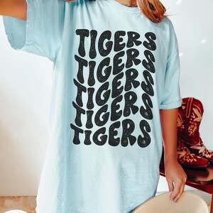 Peut inclure: Un t-shirt bleu clair avec un imprim&eacute; graphique du mot "TIGERS" r&eacute;p&eacute;t&eacute; dans une police de caract&egrave;res r&eacute;tro.