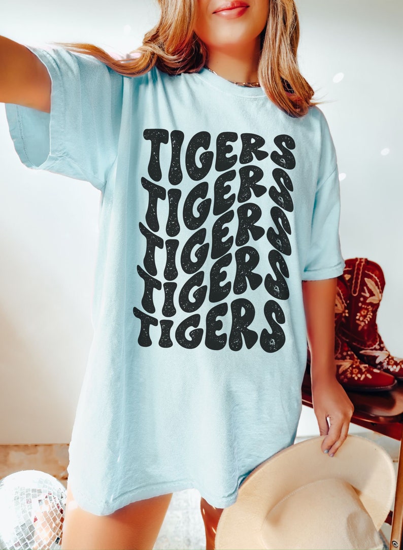 Peut inclure: Un t-shirt bleu clair avec un imprim&eacute; graphique du mot "TIGERS" r&eacute;p&eacute;t&eacute; dans une police de caract&egrave;res r&eacute;tro.