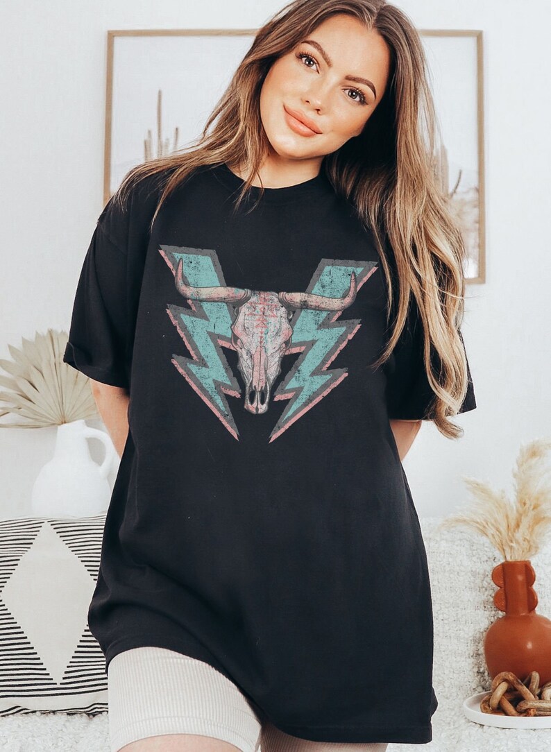 May include: Black oversized t-shirt with a graphic of a skull with horns and lightning bolts in pink, blue, and grey.