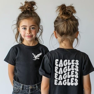 Może przedstawiać: Dwoje dzieci w czarnych koszulkach z białymi wzorami. Koszulka z przodu ma biały wz&oacute;r orła. Koszulka z tyłu ma napis "Eagles" powt&oacute;rzony cztery razy w retro czcionce.