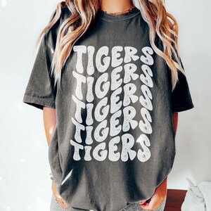 Peut inclure: Un t-shirt surdimensionn&eacute; gris fonc&eacute; avec un imprim&eacute; graphique blanc du mot "TIGERS" r&eacute;p&eacute;t&eacute; dans un style r&eacute;tro.