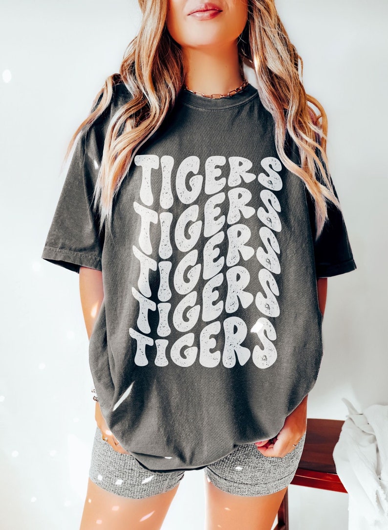 Peut inclure: Un t-shirt surdimensionn&eacute; gris fonc&eacute; avec un imprim&eacute; graphique blanc du mot "TIGERS" r&eacute;p&eacute;t&eacute; dans un style r&eacute;tro.