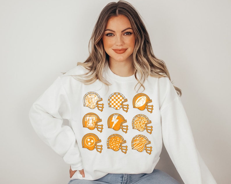 May include: White sweatshirt featuring a graphic of nine orange football helmets with various patterns, including a chequered design, a lightning bolt, and a star.
