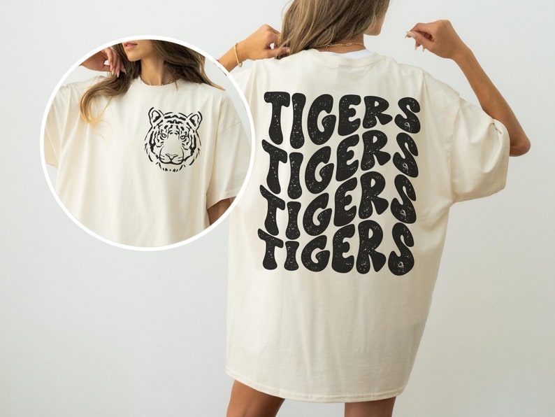以下が含まれることがあります： クリーム色のTシャツ。前面に黒い虎の顔、背面にレトロなフォントで「TIGERS」という文字が繰り返し書かれている。