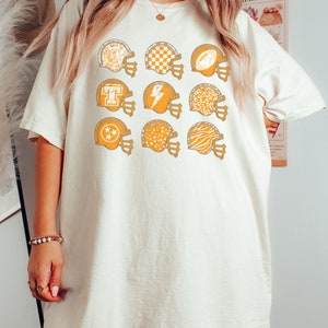 May include: A white t-shirt with a graphic of multiple orange football helmets with different patterns. The patterns include stars, checkers, animal print, and a lightning bolt.