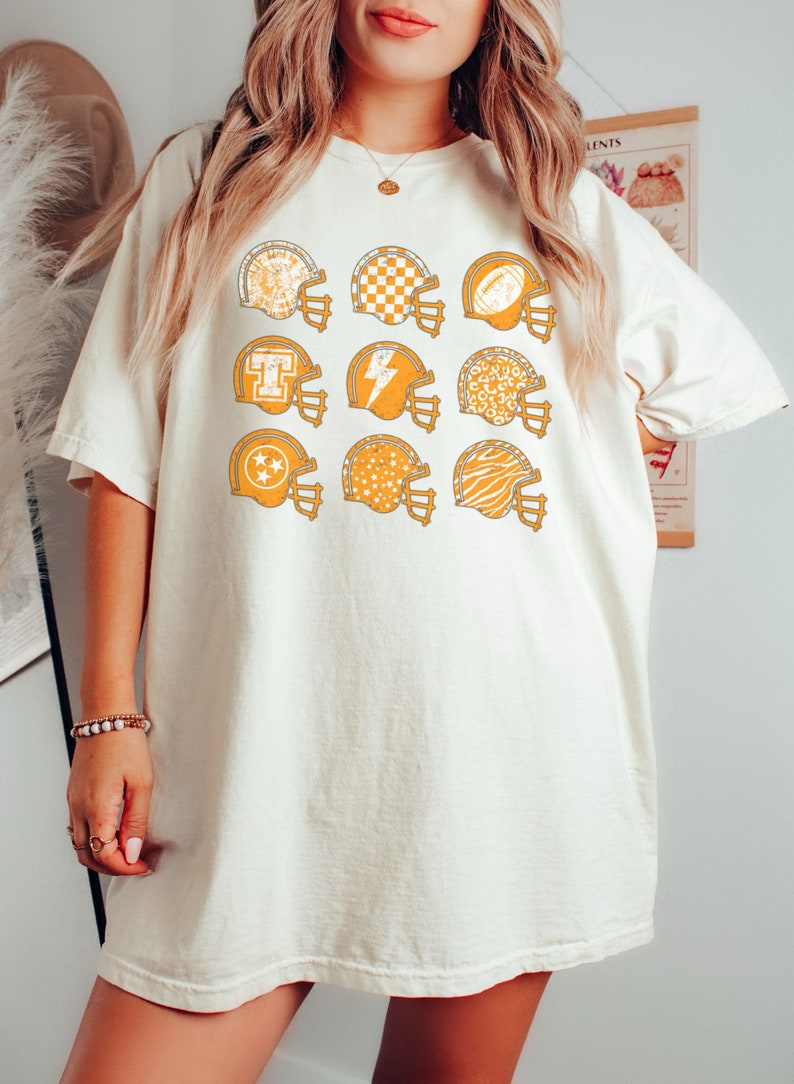 May include: A white t-shirt with a graphic of multiple orange football helmets with different patterns. The patterns include stars, checkers, animal print, and a lightning bolt.