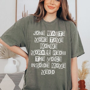 May include: A green t-shirt with a white graphic print that reads "Jone Waste Yore Toye Monme Yore All Redii The Voice Insoide Moye Yedd"