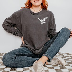 Peut inclure: Une femme portant un t-shirt &agrave; manches longues gris fonc&eacute; avec un motif d'aigle blanc sur le devant. Elle porte un jean bleu et des baskets blanches. Elle est assise sur un sol &agrave; carreaux.