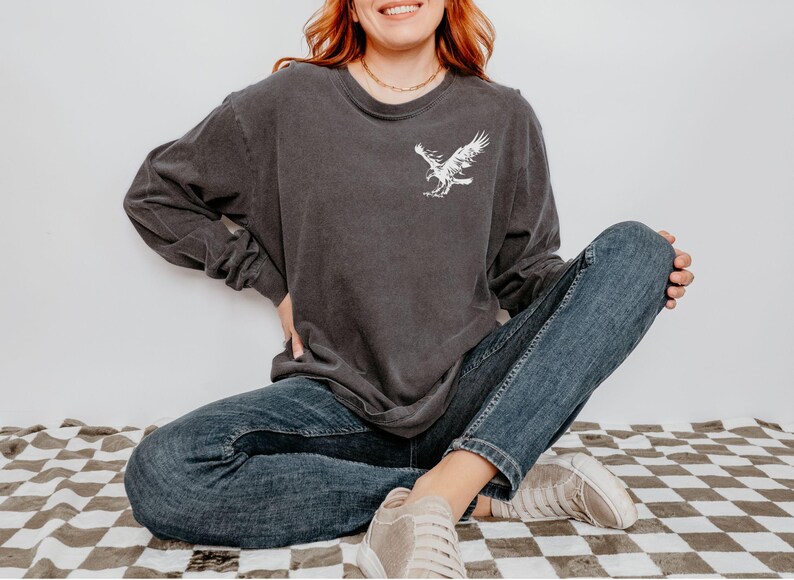Peut inclure: Une femme portant un t-shirt &agrave; manches longues gris fonc&eacute; avec un motif d'aigle blanc sur le devant. Elle porte un jean bleu et des baskets blanches. Elle est assise sur un sol &agrave; carreaux.