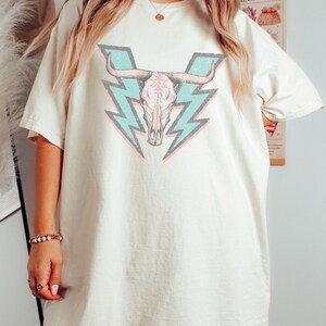 May include: A white oversized t-shirt with a graphic of a pink and white bull skull with blue lightning bolts behind it.
