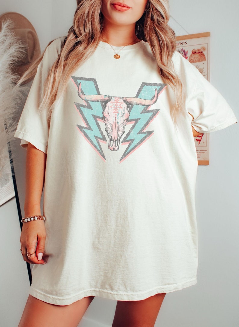 May include: A white oversized t-shirt with a graphic of a pink and white bull skull with blue lightning bolts behind it.