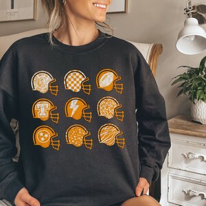 May include: Black sweatshirt featuring a pattern of orange football helmets with various designs. The helmets include patterns such as stars, chequered, and animal print.