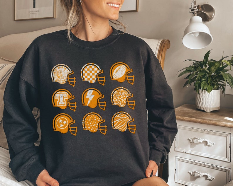 May include: Black sweatshirt featuring a pattern of orange football helmets with various designs. The helmets include patterns such as stars, chequered, and animal print.