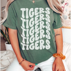 Peut inclure: Un t-shirt vert avec un imprim&eacute; graphique blanc du mot "TIGERS" r&eacute;p&eacute;t&eacute; en un motif ondul&eacute; empil&eacute;.