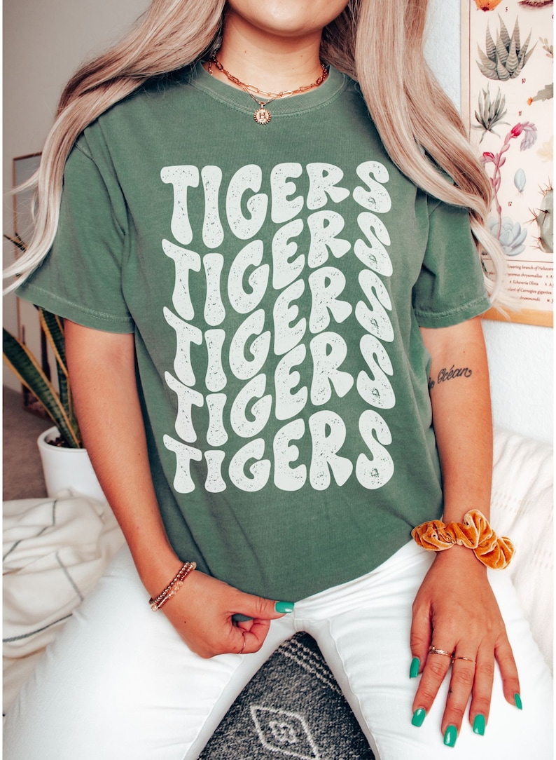 Peut inclure: Un t-shirt vert avec un imprim&eacute; graphique blanc du mot "TIGERS" r&eacute;p&eacute;t&eacute; en un motif ondul&eacute; empil&eacute;.