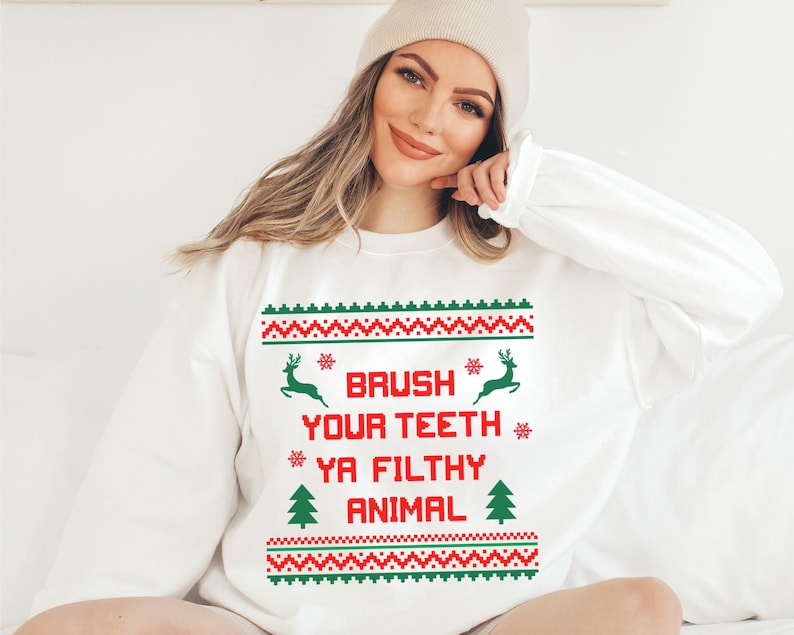 Może przedstawiać: Biały świąteczny sweter z czerwono-zielonym wzorem. Sweter ma renifera, płatki śniegu i choinkę. Tekst na swetrze brzmi "Brush Your Teeth Ya Filthy Animal".
