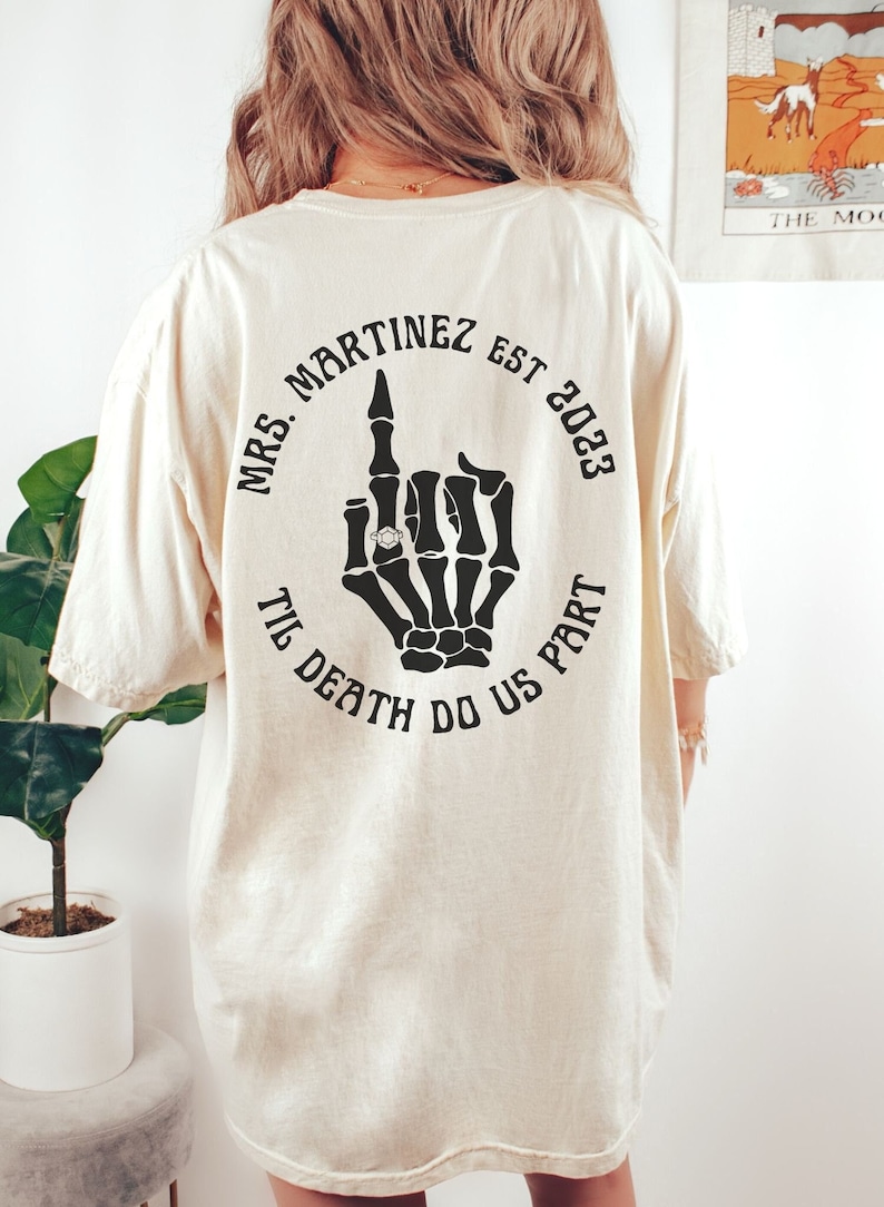 Op de afbeelding: Een wit T-shirt met een zwarte cirkelvormige afbeelding op de rug. De afbeelding toont een skelet hand met een ring aan de middelvinger, de tekst "Mrs. Martinez Est. 2023 Til Death Do Us Part" en een schedel met gekruiste beenderen.