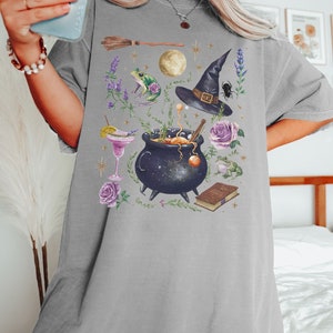 May include: A gray t-shirt with a graphic print of a witch's cauldron, a witch's hat, a frog, a broom, a book, and flowers. The graphic is in a watercolor style.