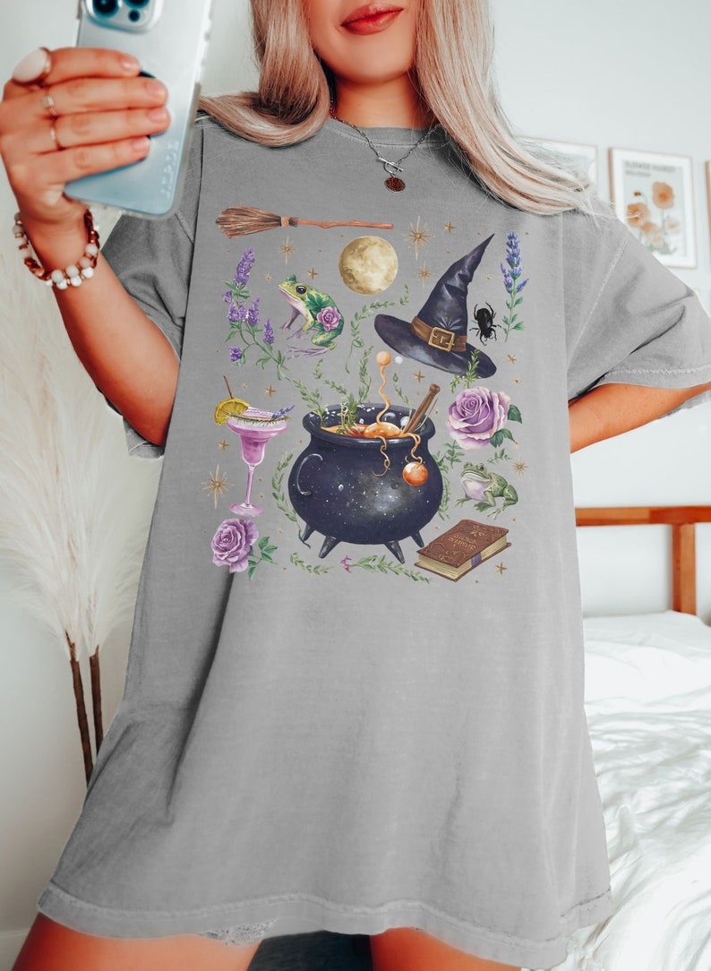 May include: A gray t-shirt with a graphic print of a witch's cauldron, a witch's hat, a frog, a broom, a book, and flowers. The graphic is in a watercolor style.
