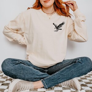 Peut inclure: Une femme portant une chemise &agrave; manches longues de couleur cr&egrave;me avec un motif d'aigle noir sur le devant. Elle est assise les jambes crois&eacute;es et porte un jean bleu et des baskets blanches.