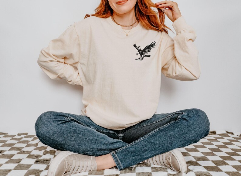 Peut inclure: Une femme portant une chemise &agrave; manches longues de couleur cr&egrave;me avec un motif d'aigle noir sur le devant. Elle est assise les jambes crois&eacute;es et porte un jean bleu et des baskets blanches.
