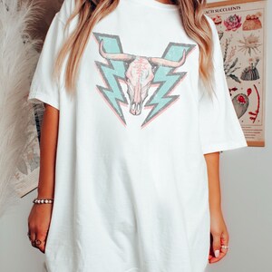 May include: White oversized t-shirt with a graphic of a turquoise and pink skull with horns and lightning bolts behind it.