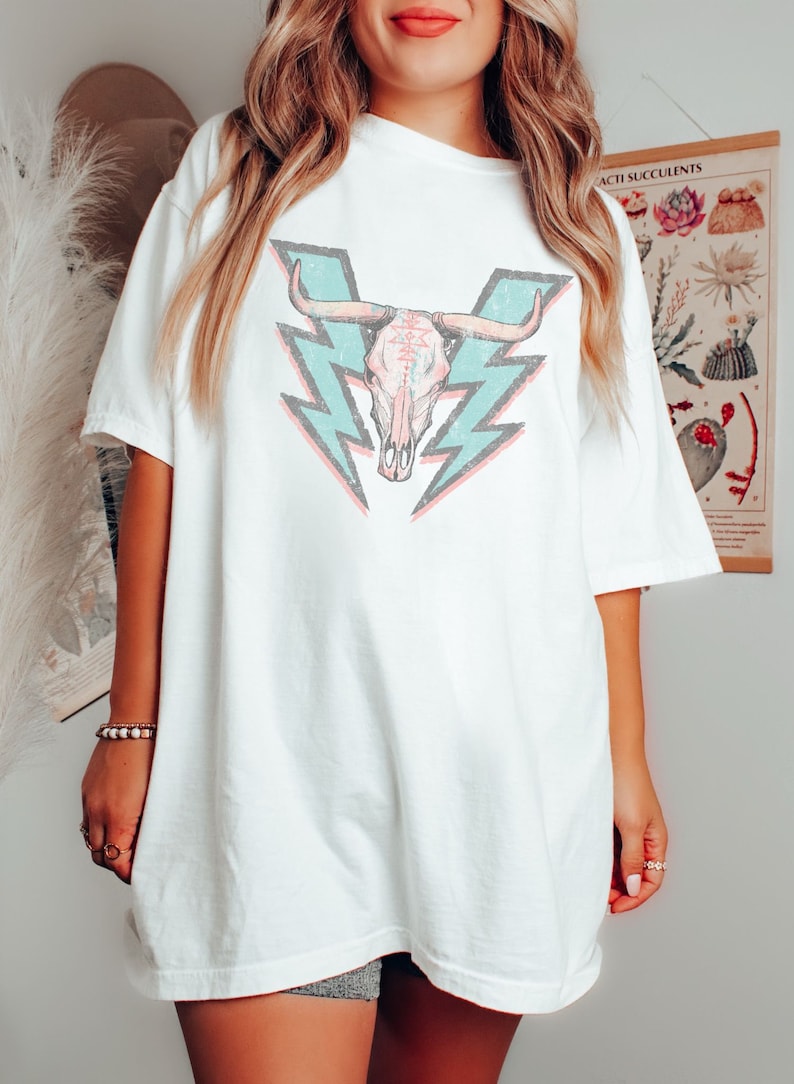 May include: White oversized t-shirt with a graphic of a turquoise and pink skull with horns and lightning bolts behind it.