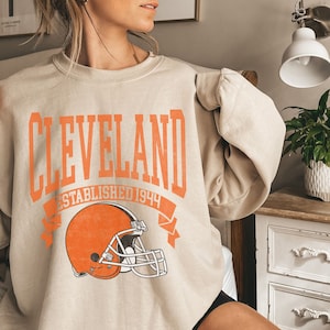 以下が含まれることがあります： オレンジ色のブロック体で「CLEVELAND」と書かれたベージュのスウェットシャツ。「ESTABLISHED 1944」とオレンジ色のフットボールヘルメットが下にあります。カジュアルなスタイルです。