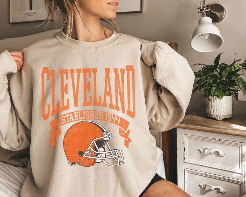 以下が含まれることがあります： オレンジ色のブロック体で「CLEVELAND」と書かれたベージュのスウェットシャツ。「ESTABLISHED 1944」とオレンジ色のフットボールヘルメットが下にあります。カジュアルなスタイルです。