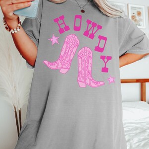 Pode incluir: Camiseta cinza oversized com um gr&aacute;fico rosa de botas de cowboy e a palavra "HOWDY" em letras rosa.