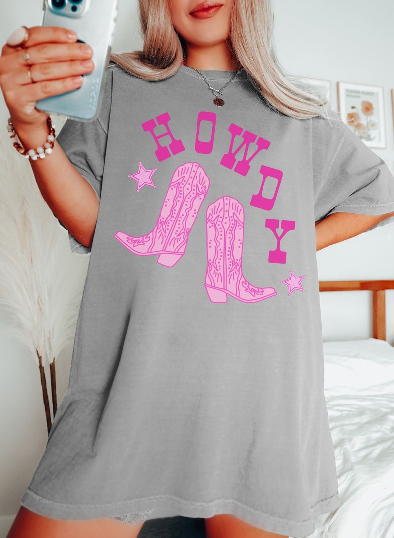 Pode incluir: Camiseta cinza oversized com um gr&aacute;fico rosa de botas de cowboy e a palavra "HOWDY" em letras rosa.
