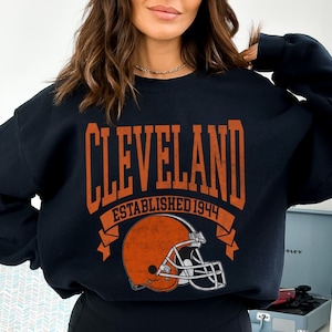 以下が含まれることがあります： オレンジ色のブロック文字で「CLEVELAND」と書かれた黒いスウェットシャツ。下に「ESTABLISHED 1944」と書かれています。オレンジ色のフットボールヘルメットのグラフィックがテキストの下にあります。スウェットシャツはクルーネックです。